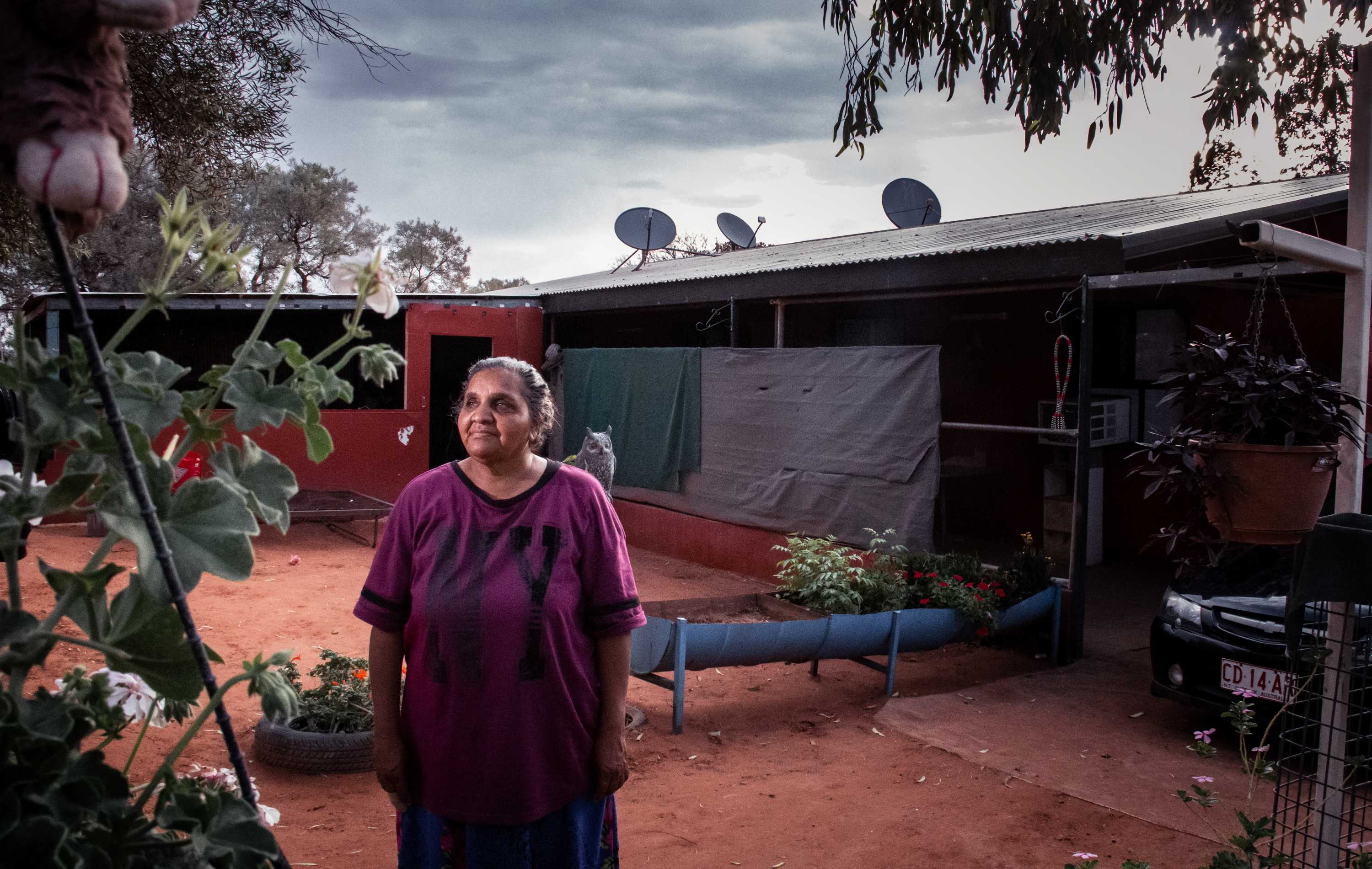 An Indigenous woman stands in front of a basic dwelling.