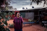 An Indigenous woman stands in front of a basic dwelling.