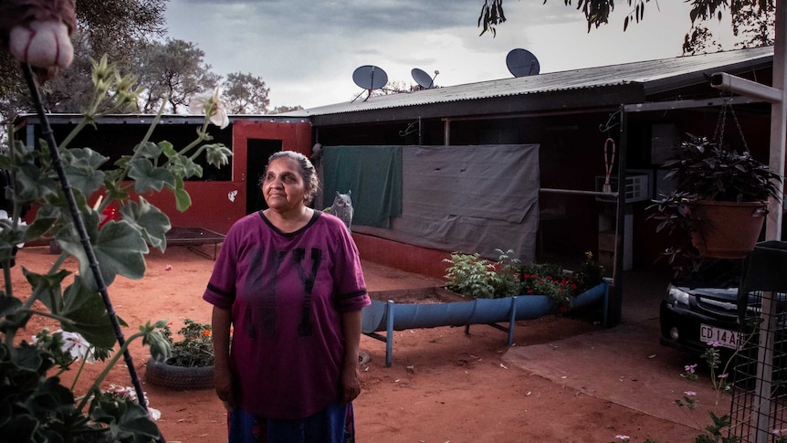 An Indigenous woman stands in front of a basic dwelling.