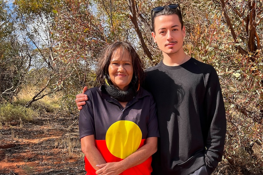 A woman wearing a tshirt with the Indigenous flag stands next to a young man in the outback