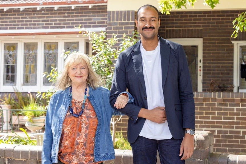 Denise Scott and Matt Okine with arms linked in front of a house, both smiling