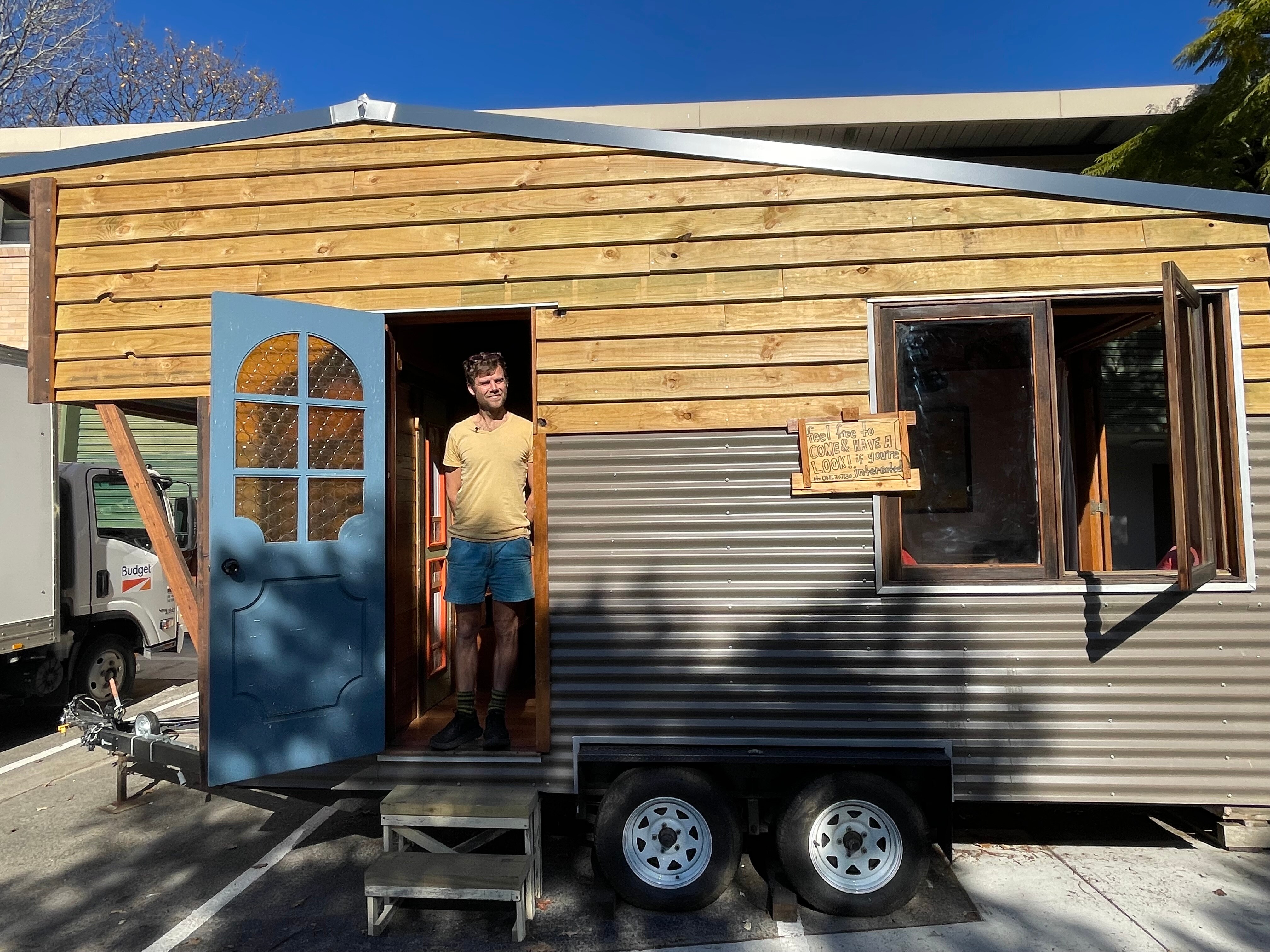A young man stands in the doorway of a tiny house with a blue door and a window.