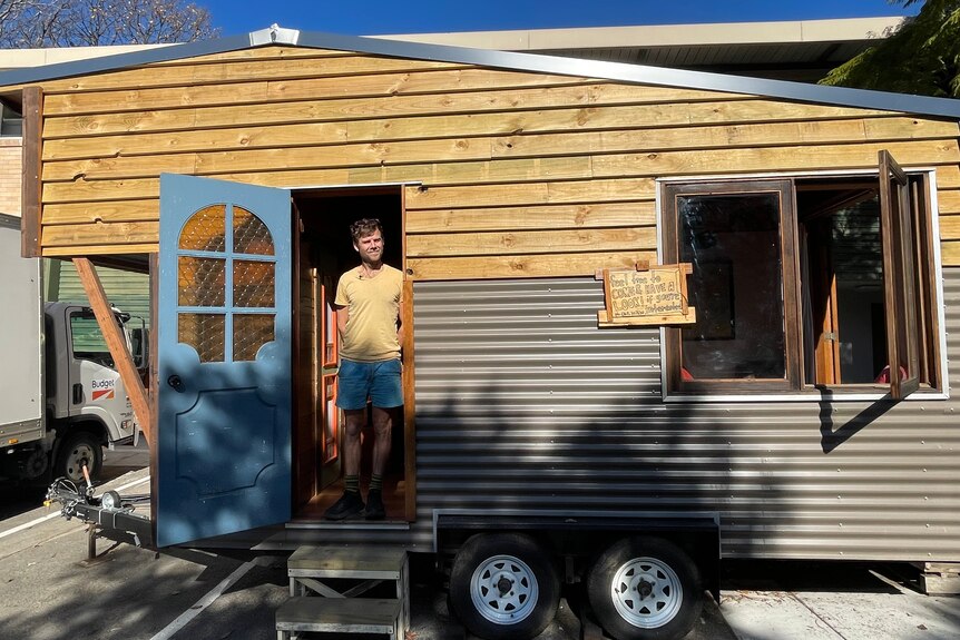 A young man stands in the doorway of a tiny house with a blue door and a window.