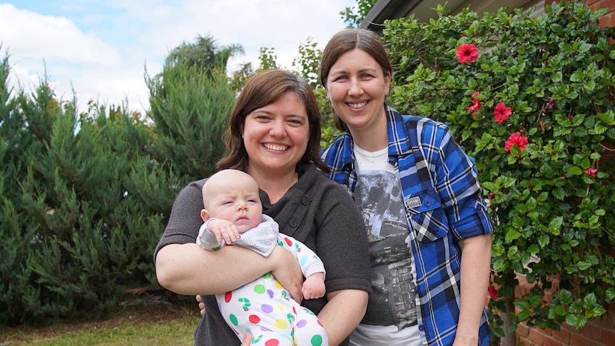 Two smiling parents holding their baby outside their red-brick home.