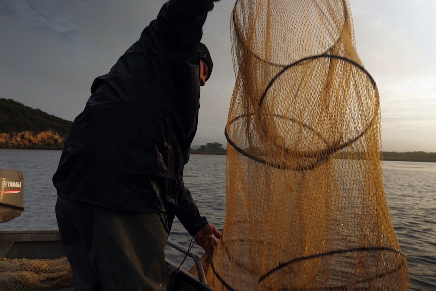 A man wearing a jacket and beanie stands in a boat and brings up his net.