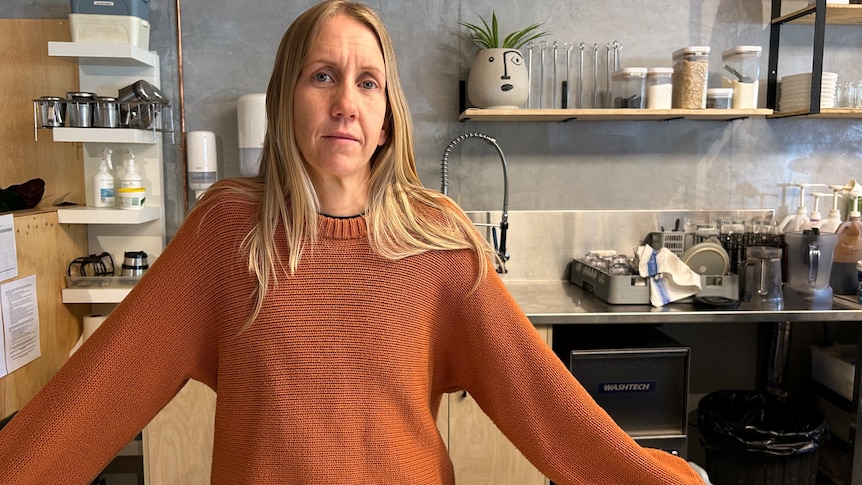 A blonde woman in an orange sweater stands behind a cafe counter top.