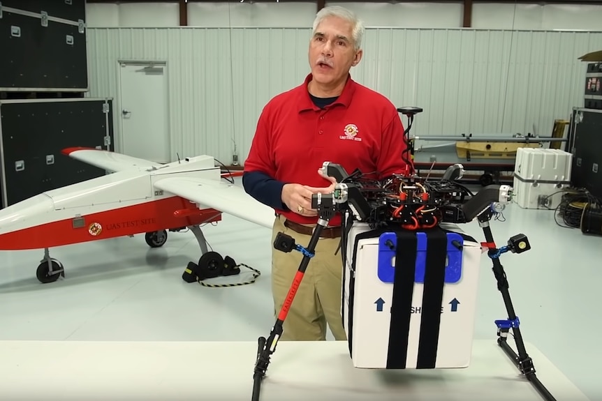 A man wearing a red shirt stands in a workshop with drones.