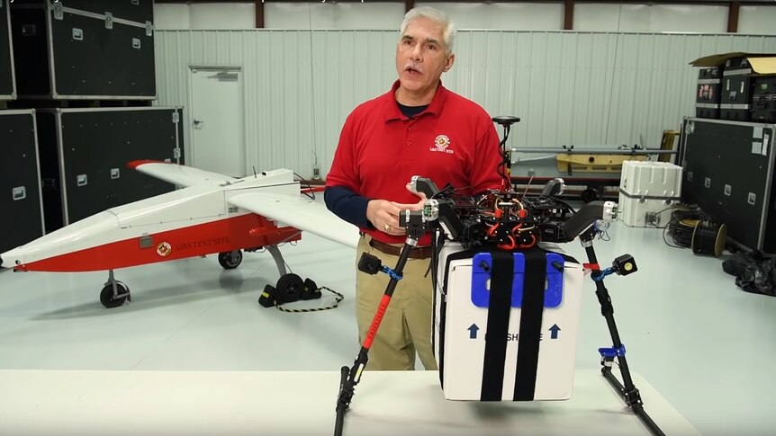 A man wearing a red shirt stands in a workshop with drones.