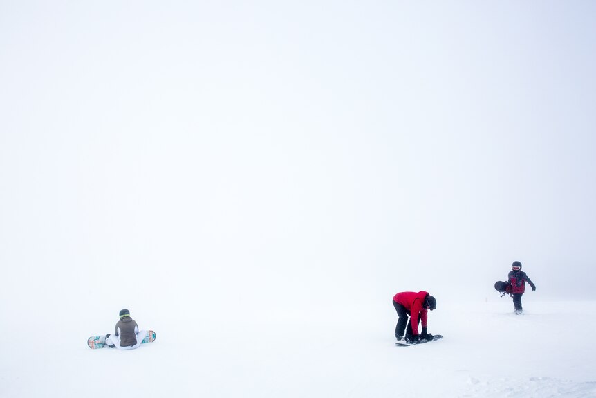 Three snowboarders, one trying to stand up, one trudging and one on the ground against a whited out landscape.