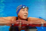 An elderly woman in a pool resting on the side wearing a blue swimming cap