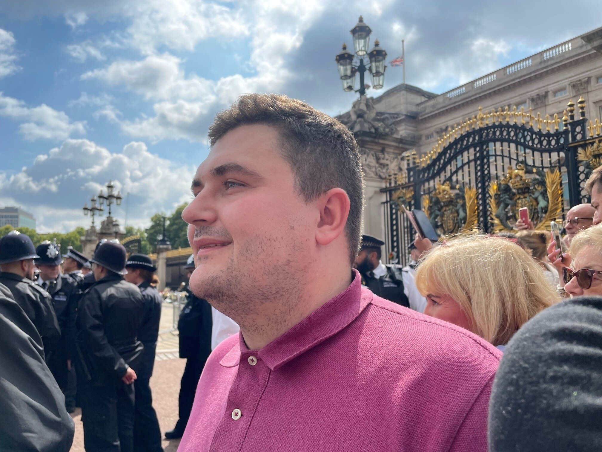 A man in a pink polo shirt looking emotional at the gates of Buckingham Palce