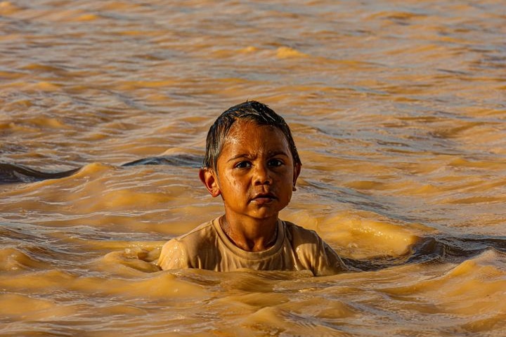 A serious Indigenous child wears a cream tee and is swimming in murky waters. 