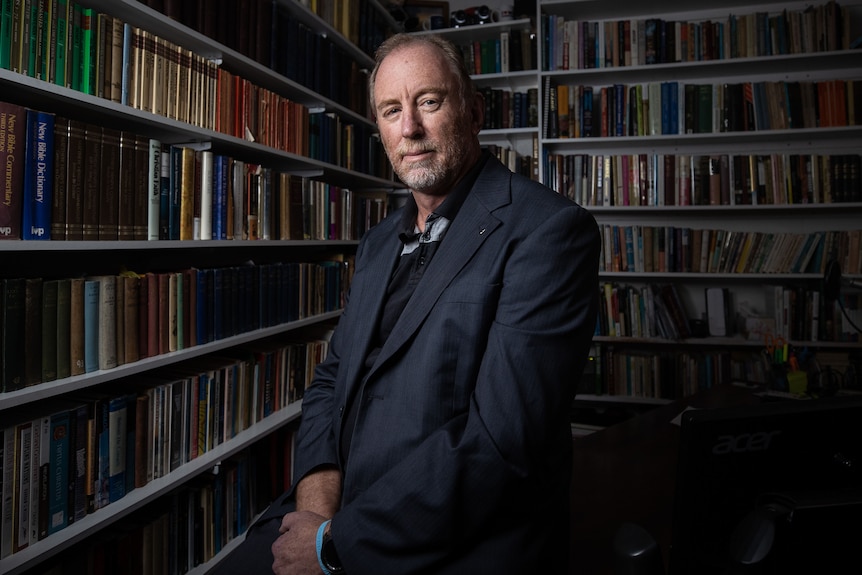 A man sits on a desk with bookcases around him.