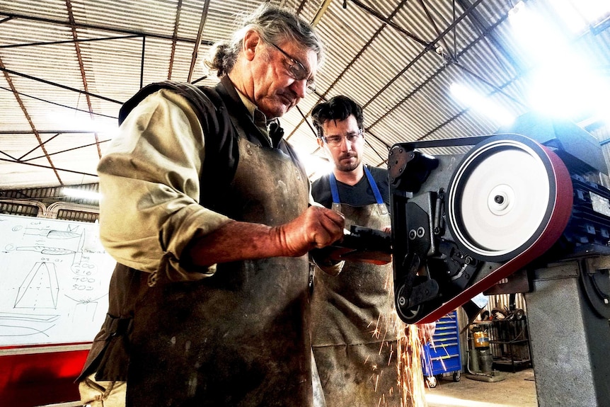 A father and a son use a machine to make a knife in a workshop.