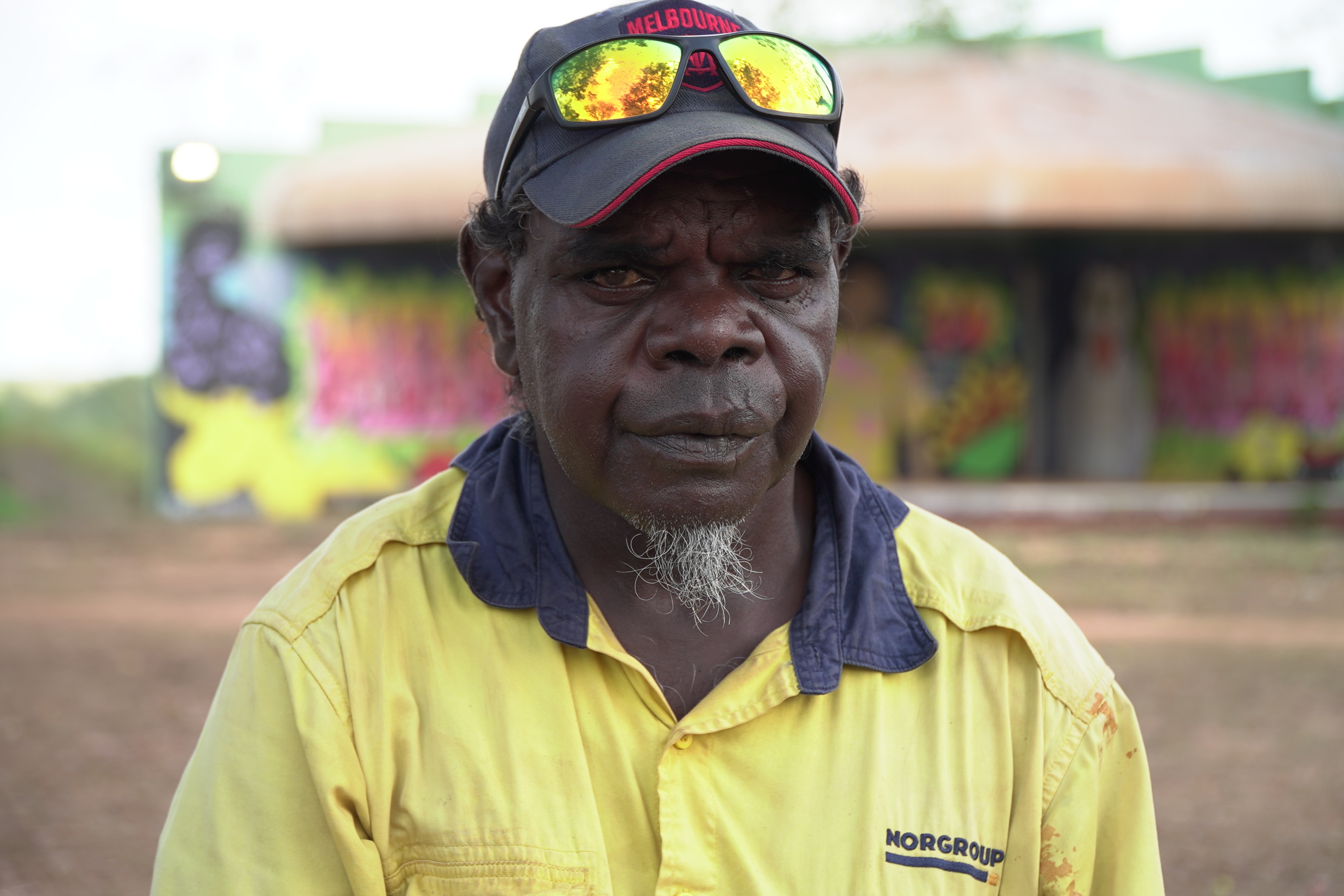 Arthur James Junior Karui looks directly at the camera while wearing a yellow, high-vis shirt and a baseball cap with sunglasses