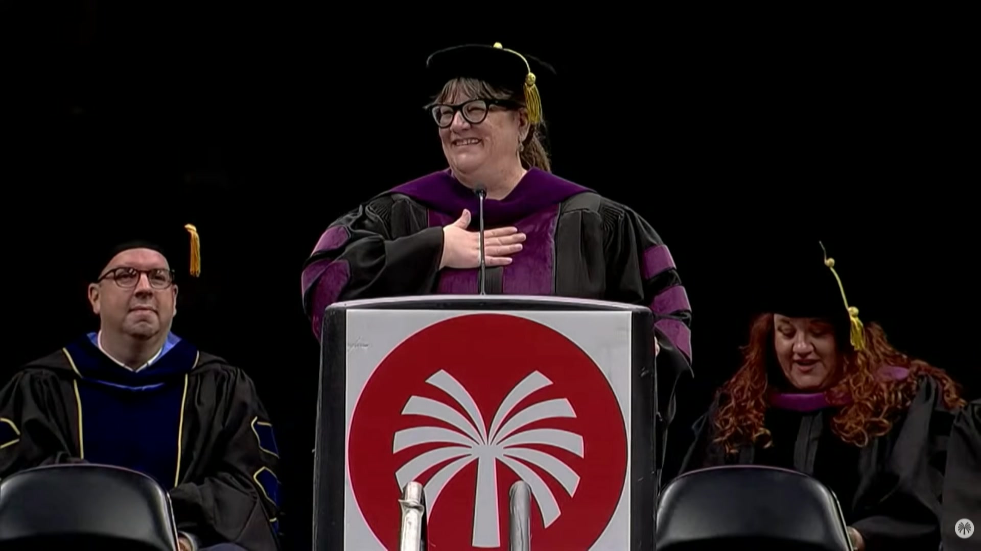 A woman at a lectern in a graduation outfit holds her hand to her chest.
