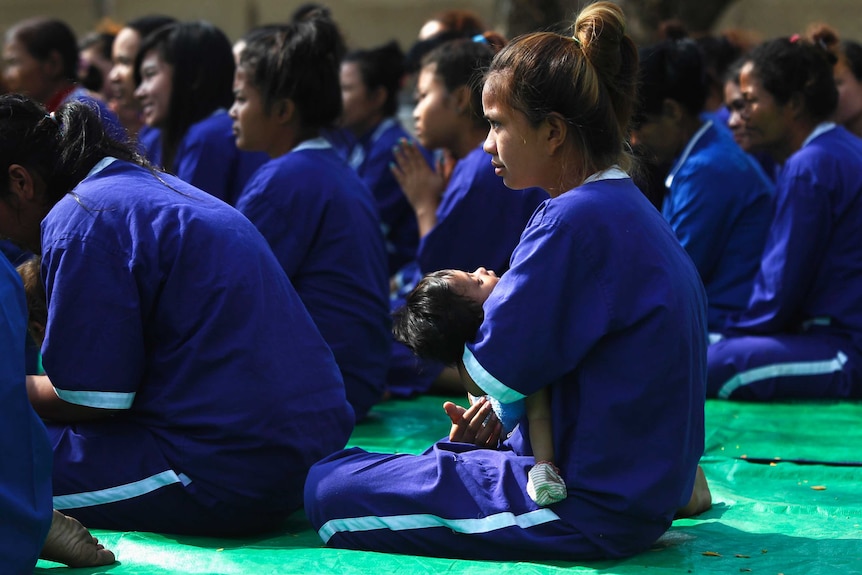 A woman in blue prison outfit with her baby sitting and looking forward.