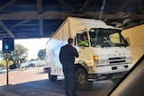 A truck wedged under a bridge, with a man standing beside.