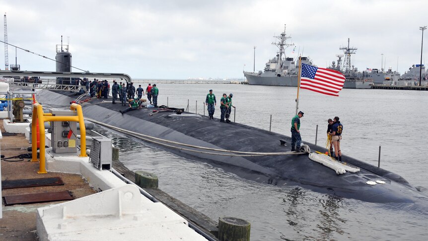 A submarine rests in a dock, crews of people walking on its surface, and an American flag planted at its bow.