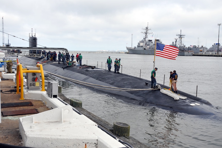 A submarine rests in a dock, crews of people walking on its surface, and an American flag planted at its bow.