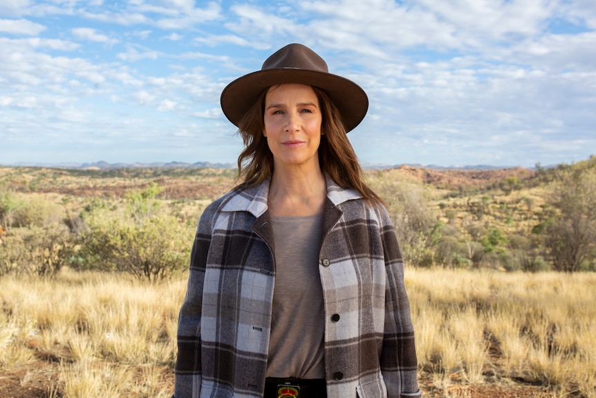 Rachel Griffiths stands in front of a grassy landscape.  She is wearing a brown hat. 