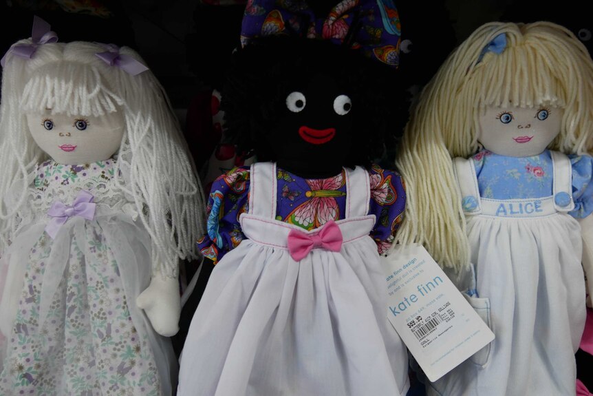 A golliwog doll in between two white rag dolls on the shelf at a shop.