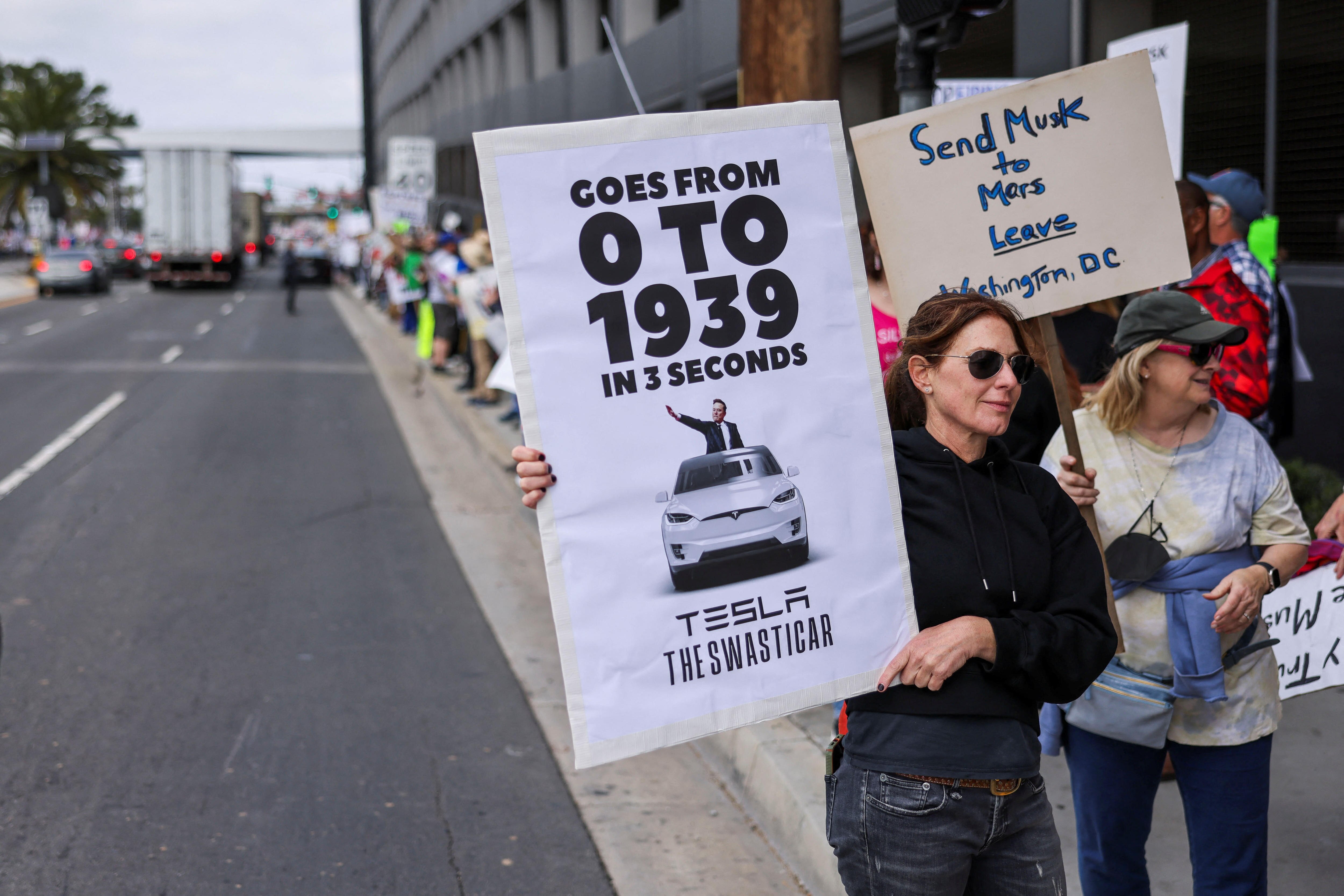 A woman holds a protest sign with Elon Musk and Tesla that reads "Goes from 0 to 1939 in 3 seconds." 