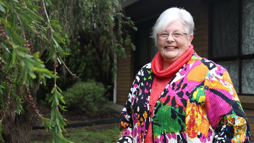 A woman in a colourful jacket smiles at the camera, a tree to the left and brick house behind her