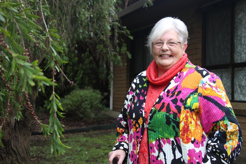 A woman in a colourful jacket smiles at the camera, a tree to the left and brick house behind her