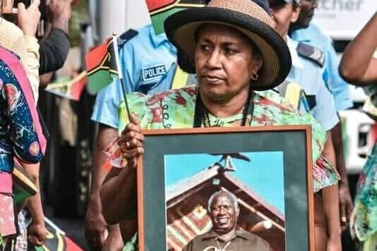 A woman marches in a parade, holding a framed photo of a man smiling.