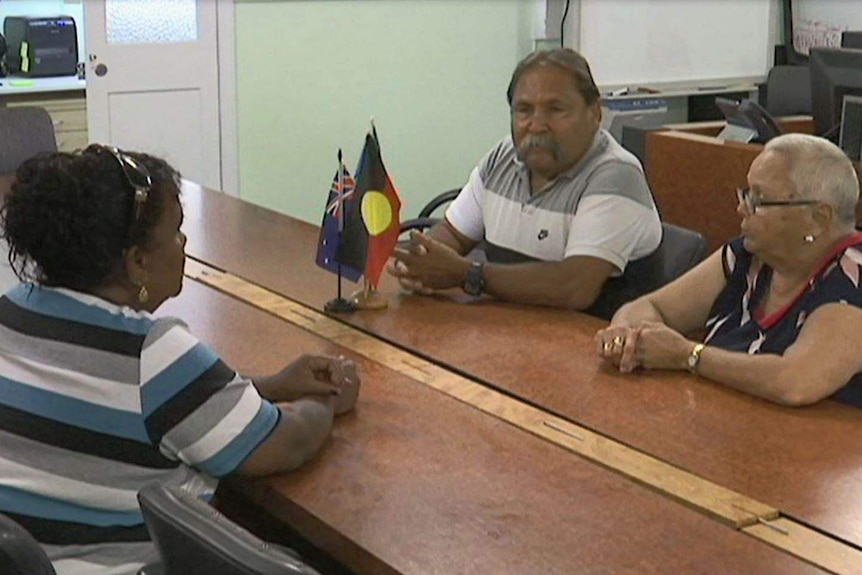 three elders sitting around a table facing eachother with aboriginal flag on the table