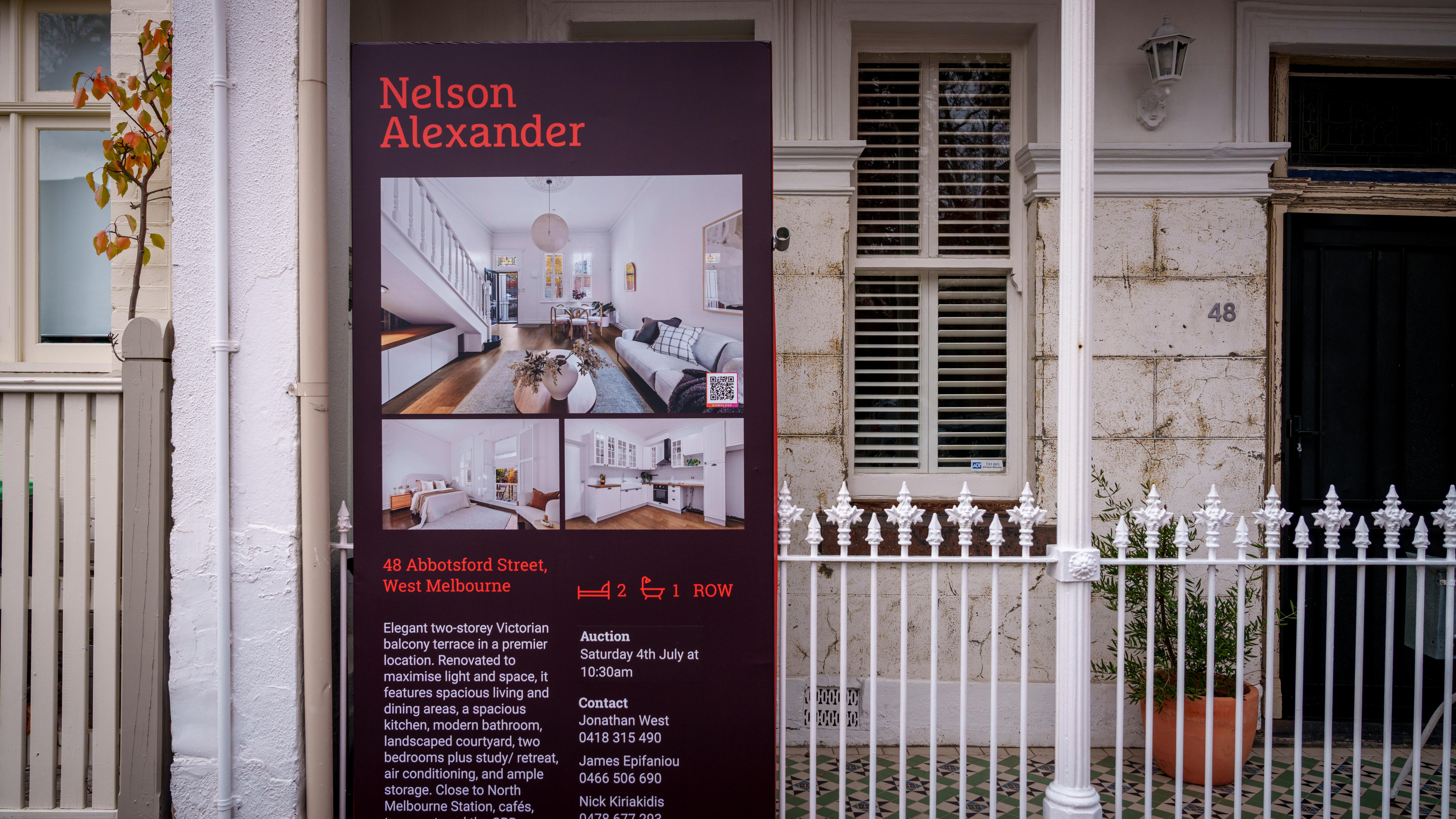A large sign with real estate information attached to a white picket fence of a white brick terrace home.