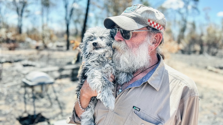 Man cuddles small grey dog in burnt out landscape.