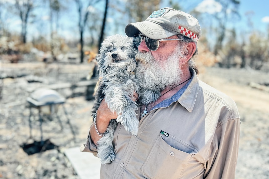 Man cuddles small grey dog in burnt out landscape.