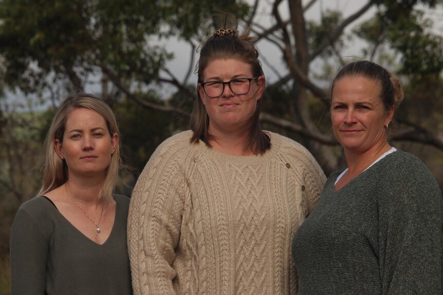 Three women stand looking into the camera, on a dirt road near some trees