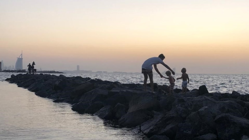 The silhouette of a man playing with two young children, with the ocean and the Dubai skyline in the background.