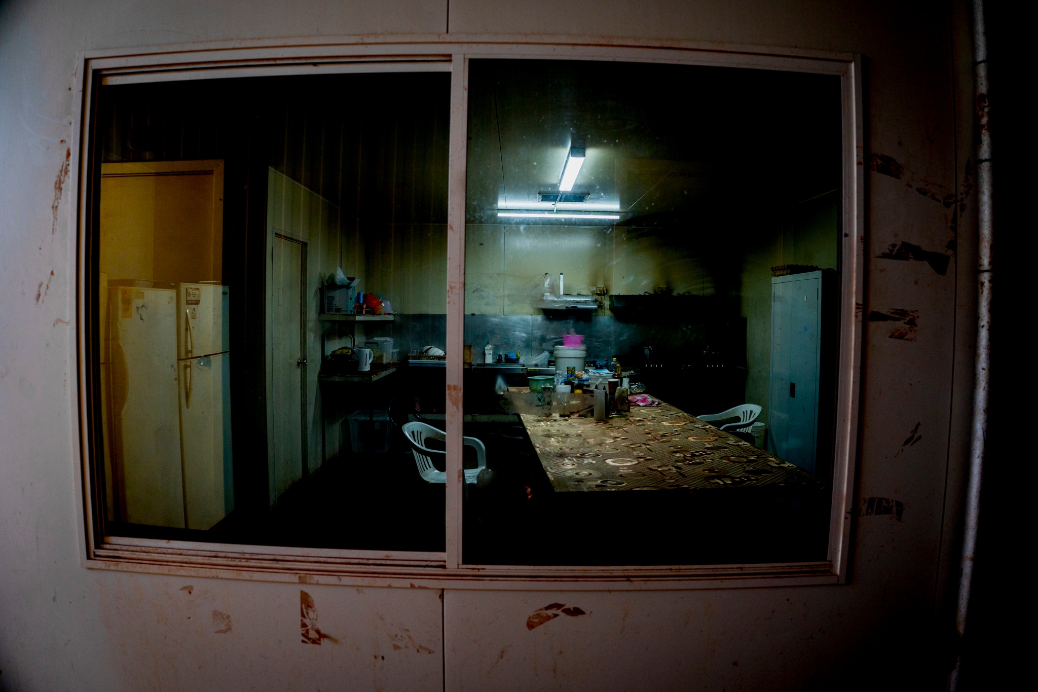 a dark, humble kitchen with plastic chairs, fluorescent lights and steam rising from a pot on the stove. 