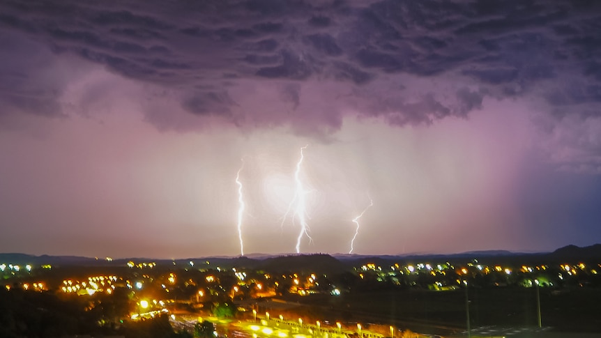 Lightning and storm clouds over a town, lit up at night. There are three forks of lightning lighting the grey cloudy sky. 