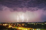 Lightning and storm clouds over a town, lit up at night. There are three forks of lightning lighting the grey cloudy sky. 