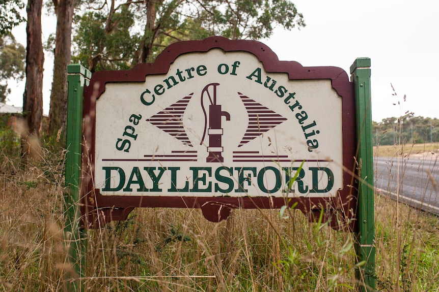 A green and maroon sign in the grass beside a road, saying Spa Centre of Australia Daylesford, with a handpump in the middle.