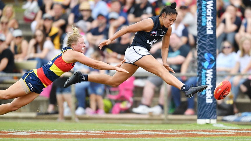 Carlton AFLW player stretches mid-air to boot the ball for a goal, beating a despairing defender.