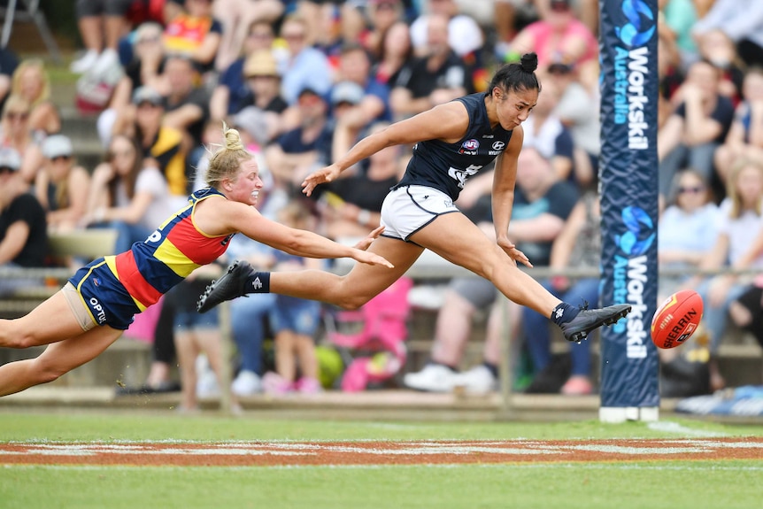 Carlton AFLW player stretches mid-air to boot the ball for a goal, beating a despairing defender.