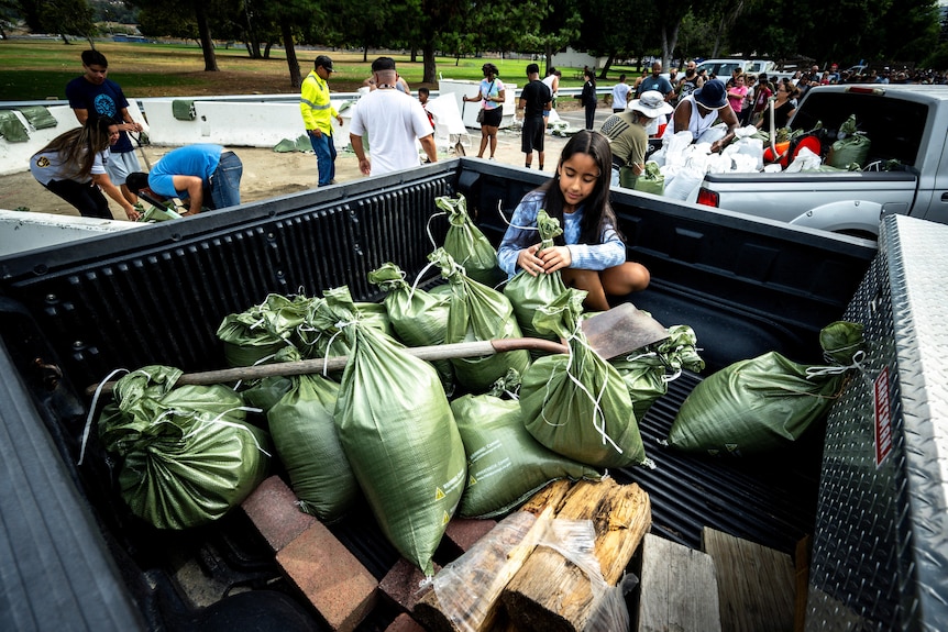 A girl with black hair sits in the tray of a ute with green sandbags