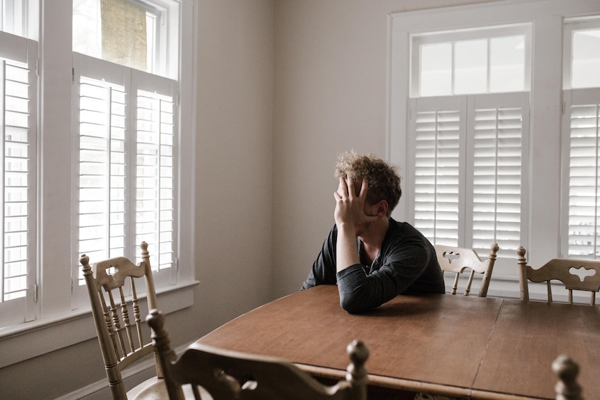 A man in a bare room seen side-on, sitting at a table leaning his head into his hand and facing towards a window.