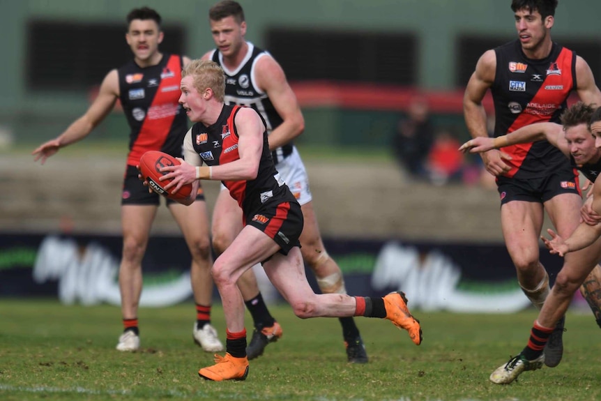 A man with blonde hair wearing a black and red football guernsey runs away from a group of players with a football in his hands
