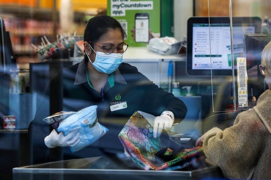 A supermarket worker wearing a face mask and gloves packs a shopper's bag