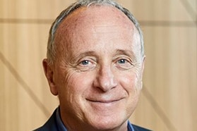 A smiling portrait of a middle aged man wearing a navy shirt and jacket.