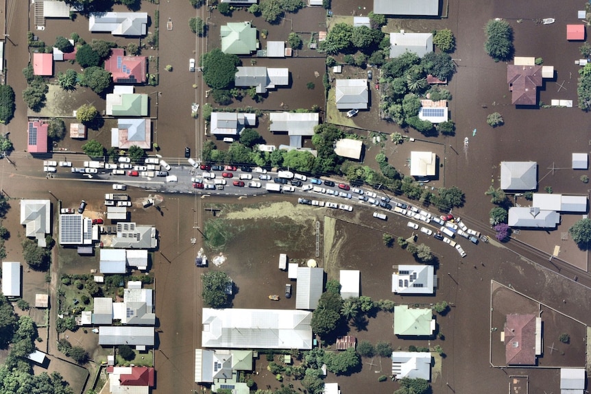 An aerial view of several dozen cars on a road, surrounded by flooded houses and shops in a town.