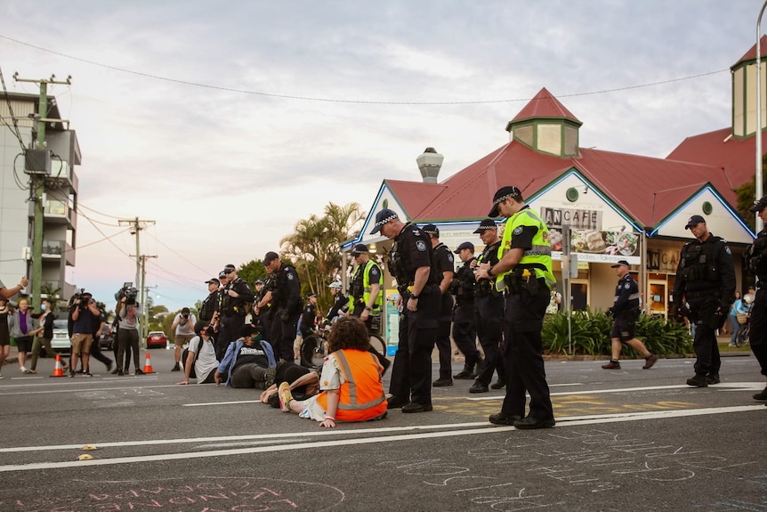 Protesters sit on a line on a chalked road as police stand over them.