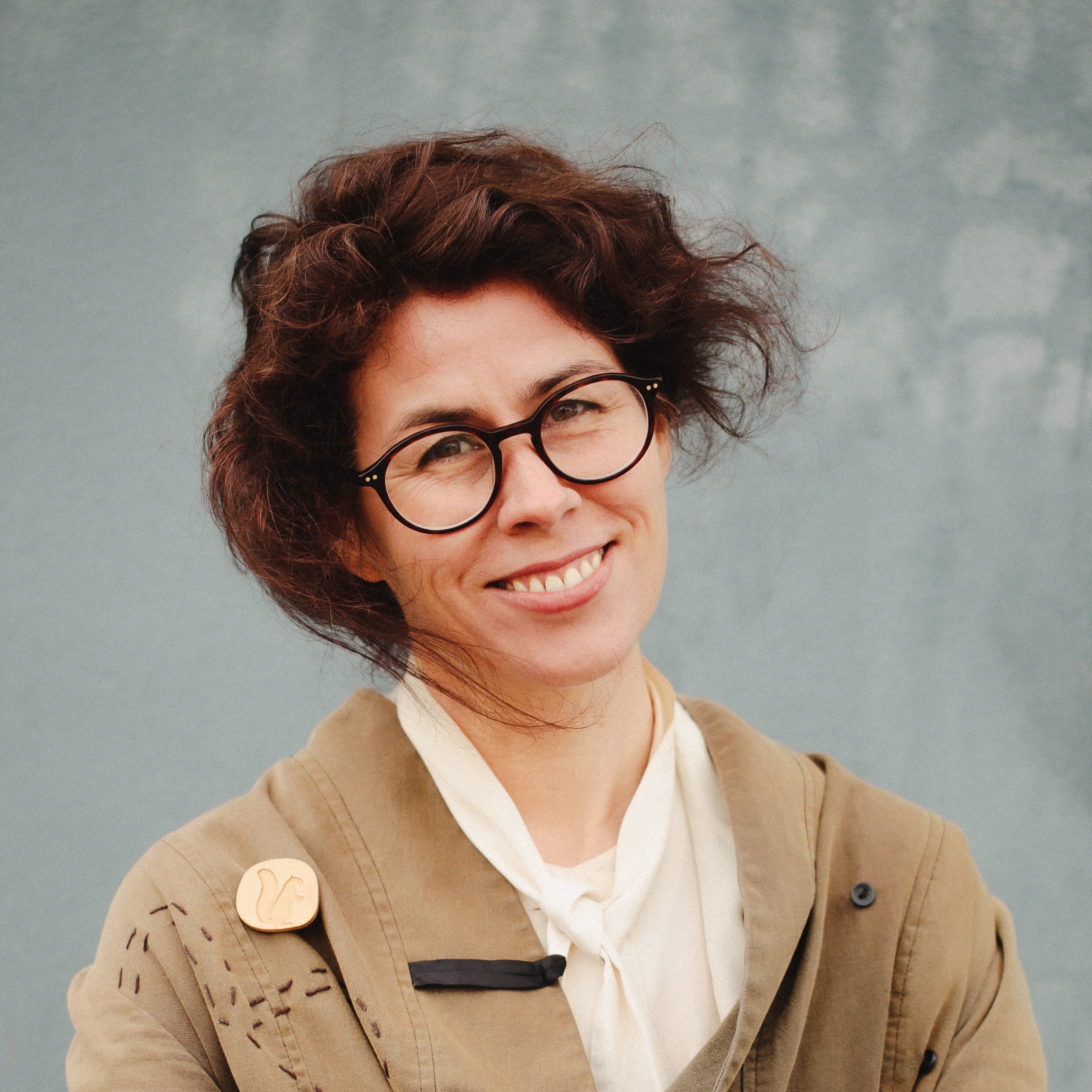 A 40-something woman with brown hair and round glasses wearing light brown cardigan, smiling at camera with head tilted.